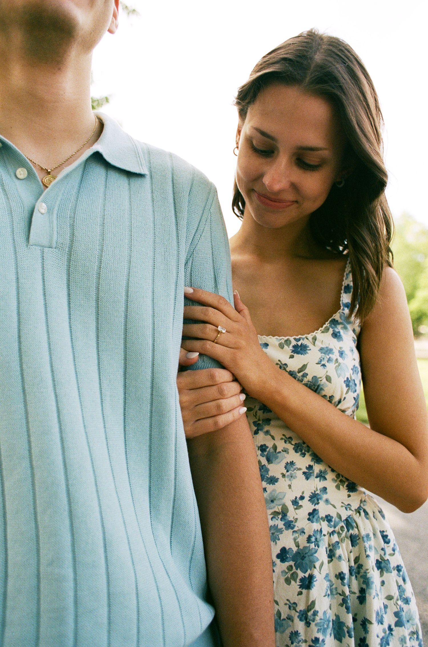 A smiling woman in a floral dress holds a man's arm, showcasing an engagement ring outdoors.