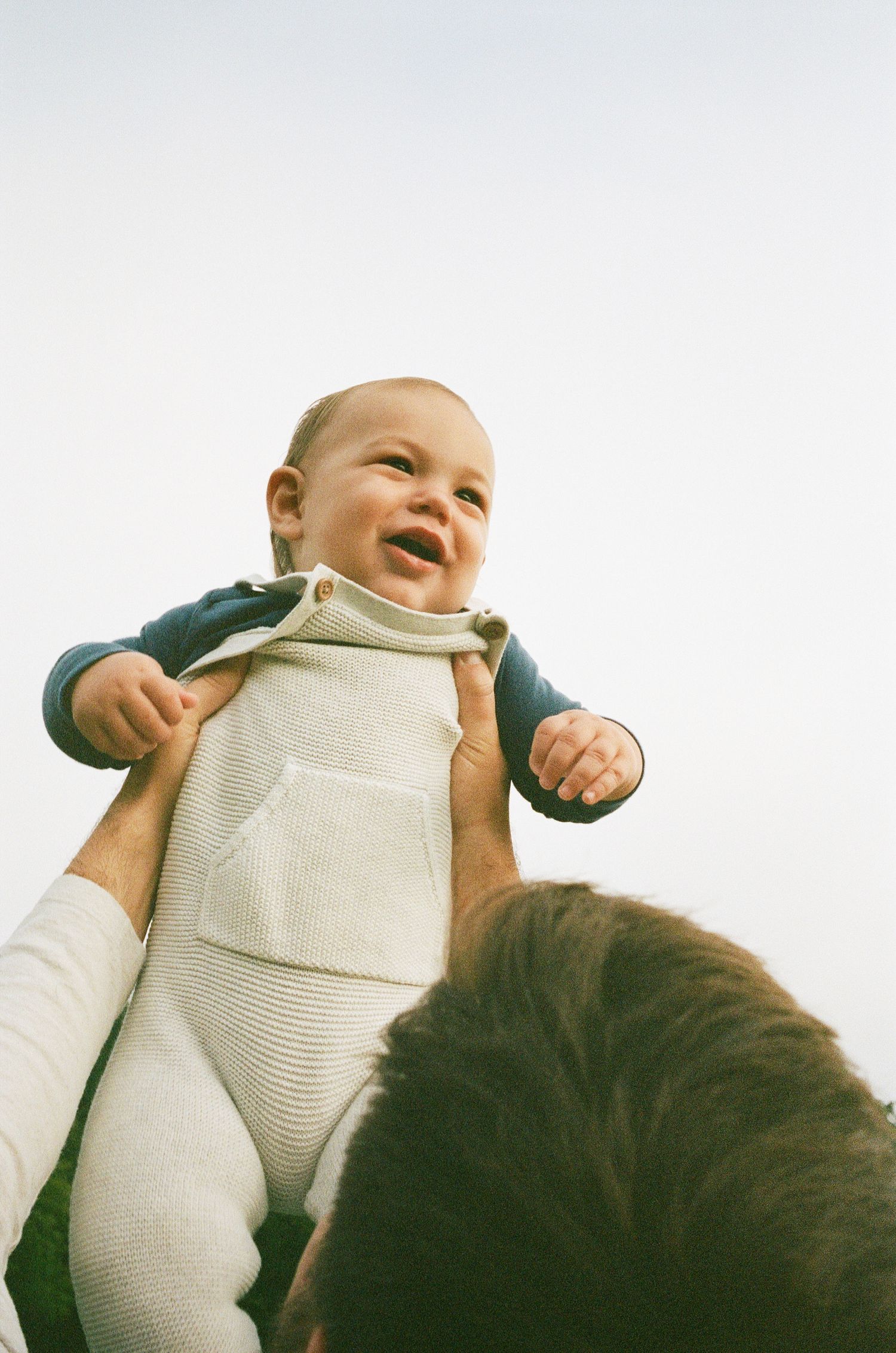 A baby in a white onesie is being lifted up and smiling joyfully while playing.