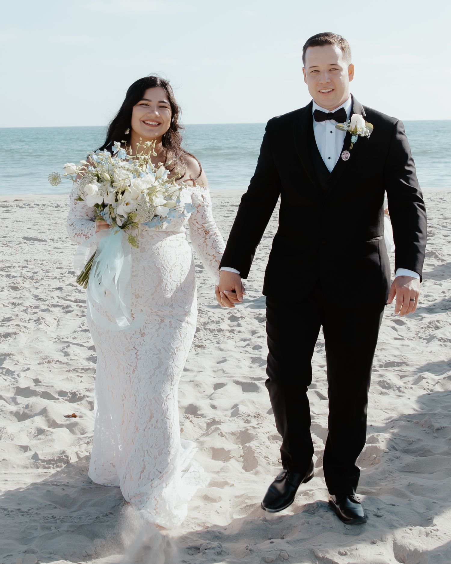 A couple walks hand in hand along the beach in wedding attire with white flowers.