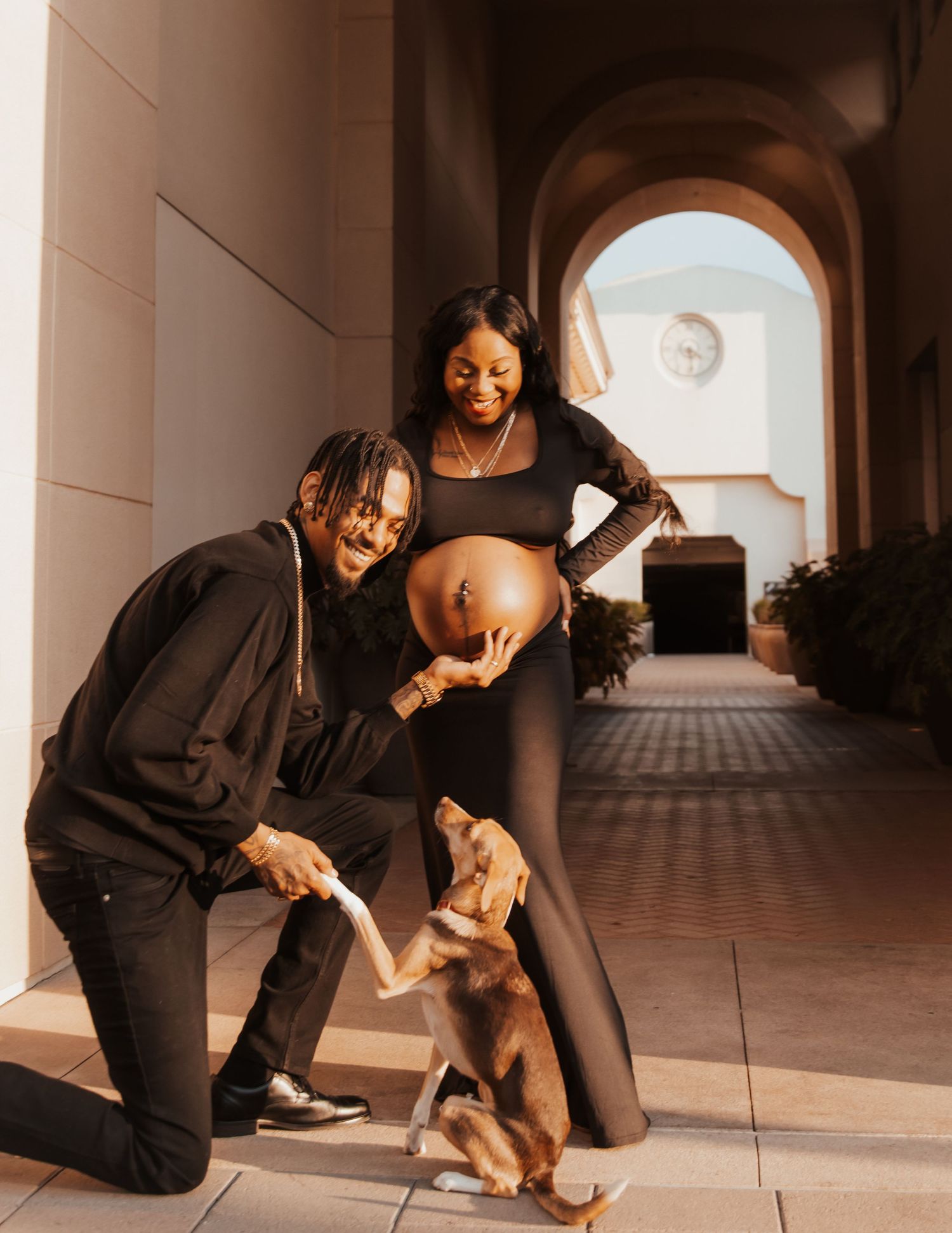 Playful couple in black outfits interacting with their dog in an arched hallway.