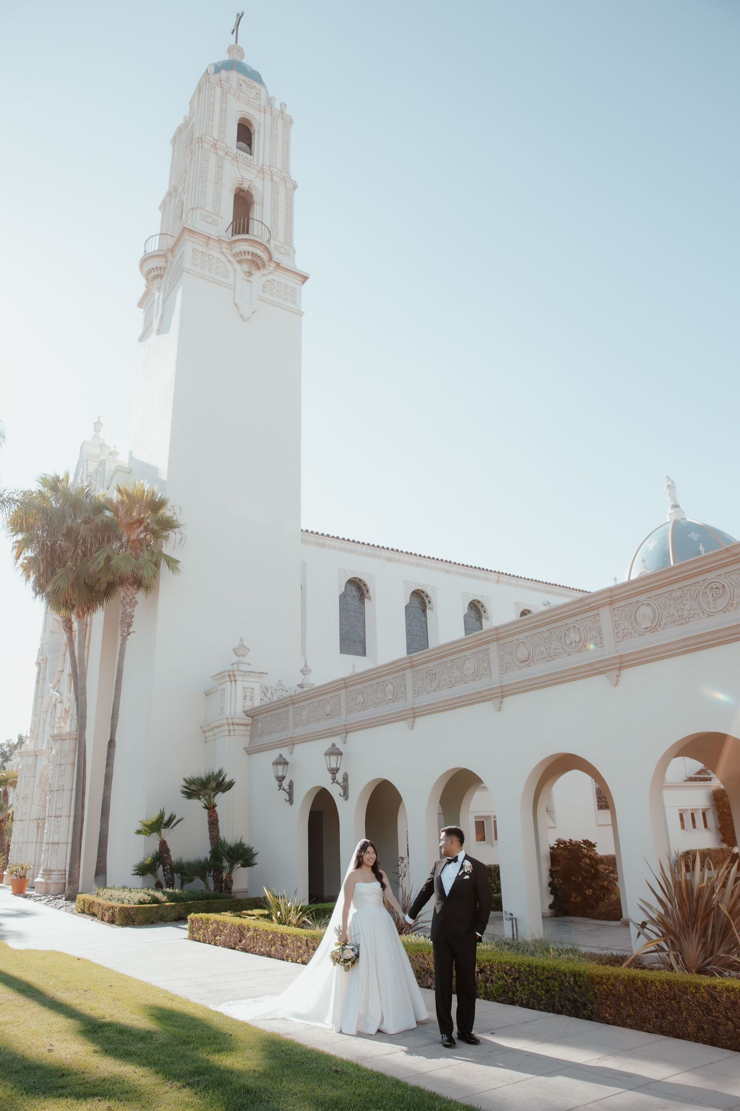A couple poses in front of the white Spanish colonial architecture of Immaculata Church at USD with its iconic bell tower.