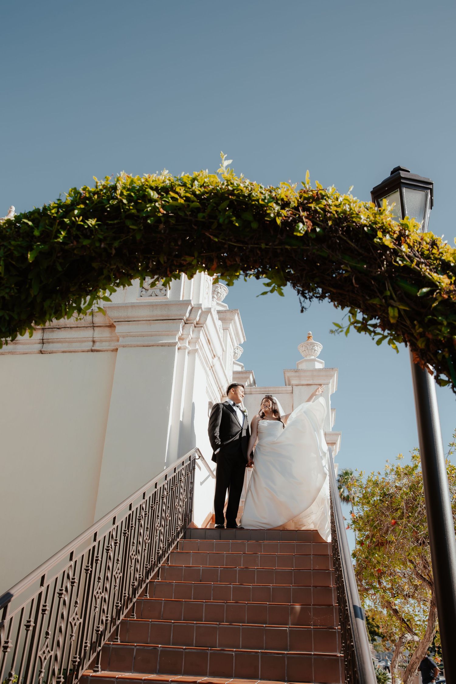 Newlyweds stand under a floral arch on white stone steps against a bright blue sky.