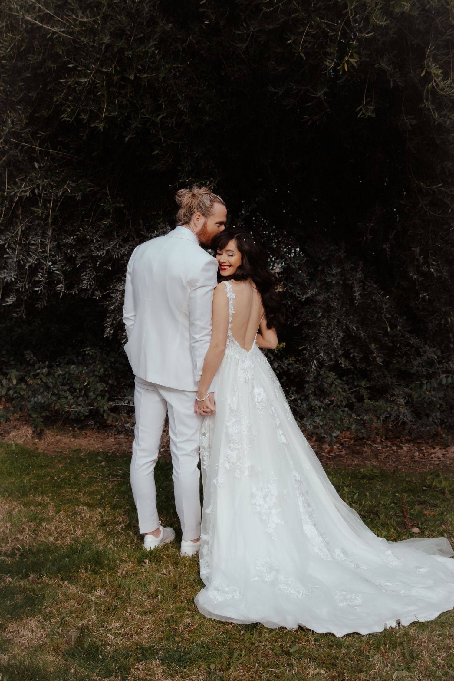 A romantic wedding couple walks together on grass wearing white attire with a flowing bridal gown train.