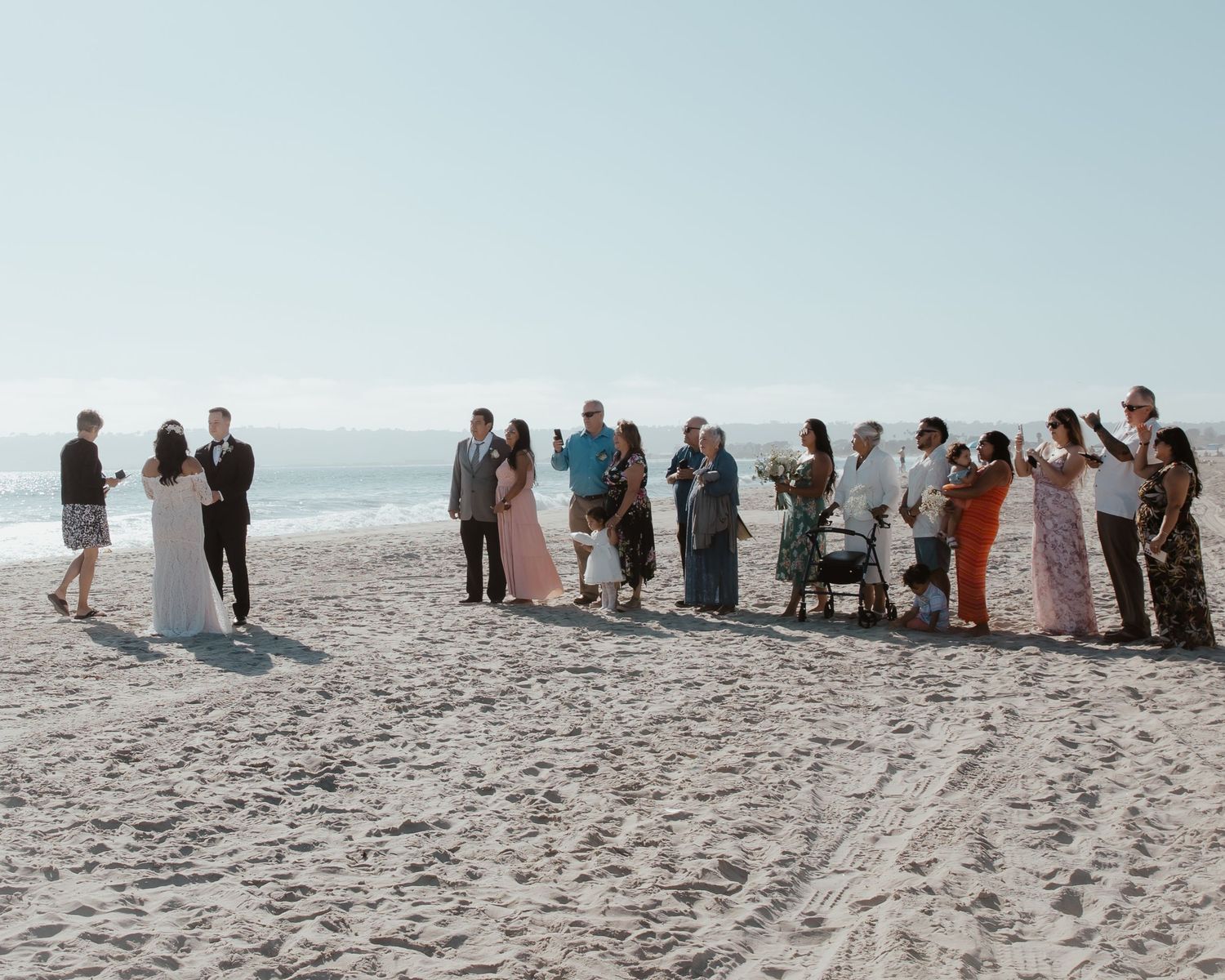 Wedding guests gather on beach to witness ceremony against ocean backdrop.