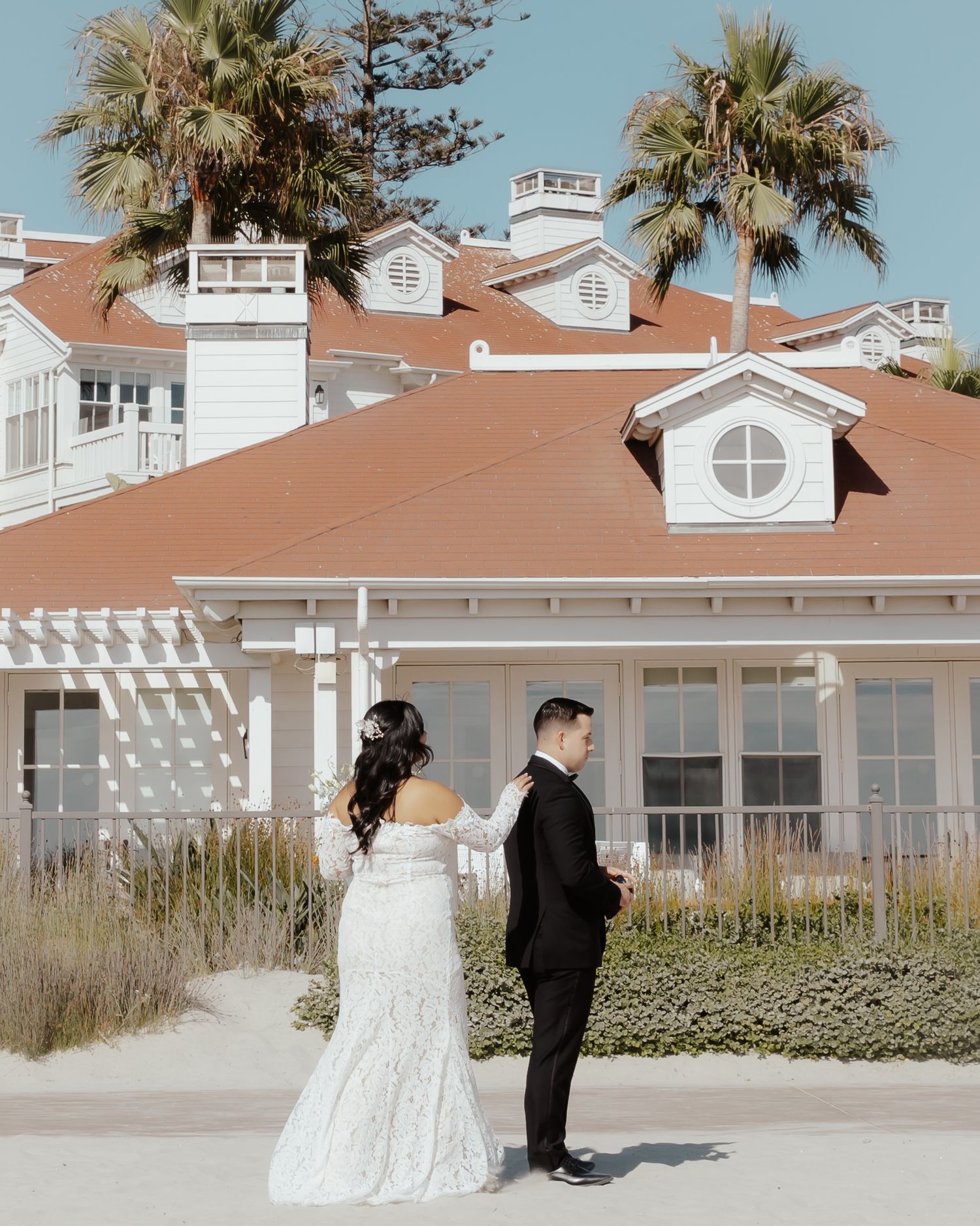 A wedding couple poses in front of a coastal resort with palm trees and white buildings.