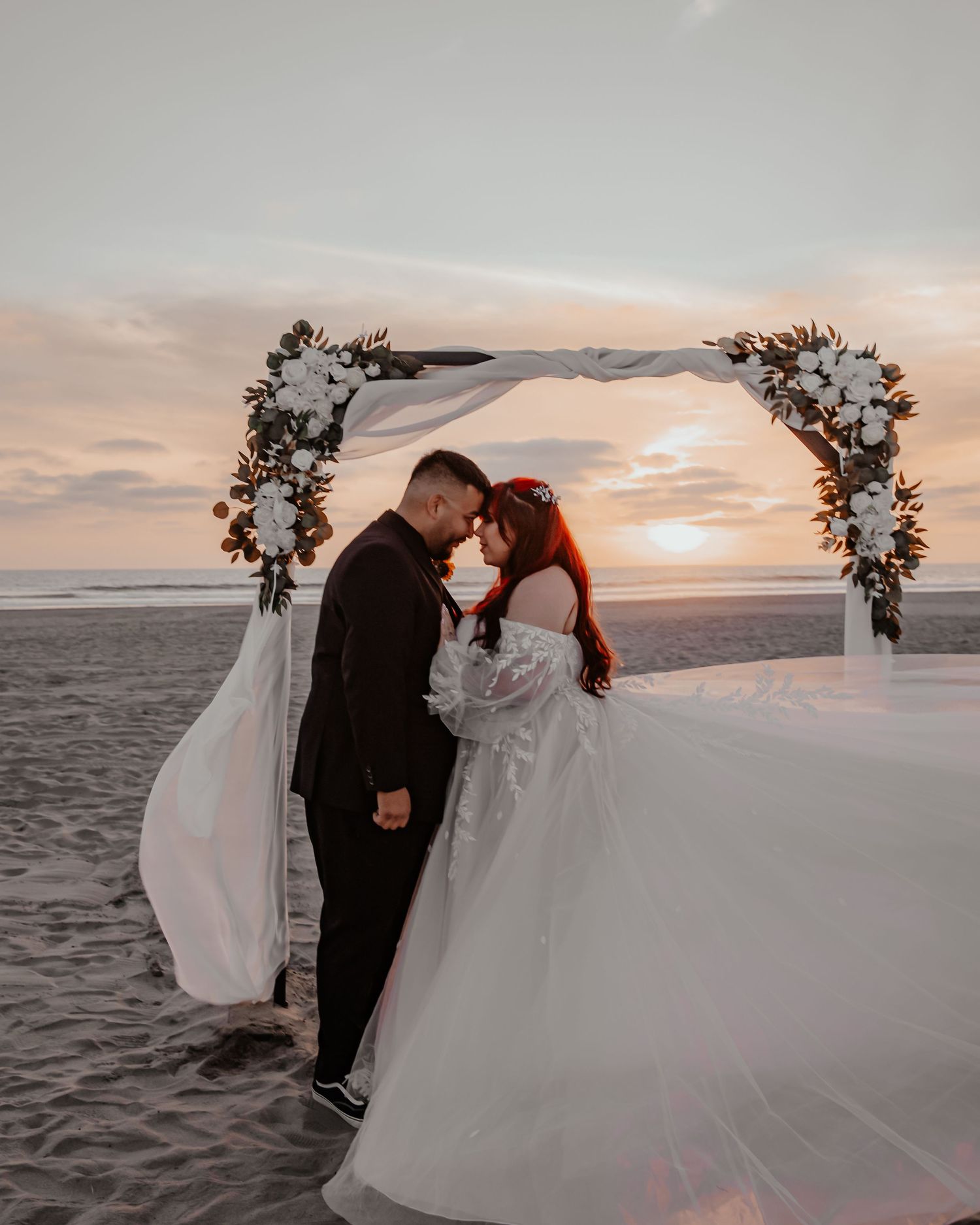 A beach wedding scene at sunset with floral arch and flowing white dress catching the breeze.