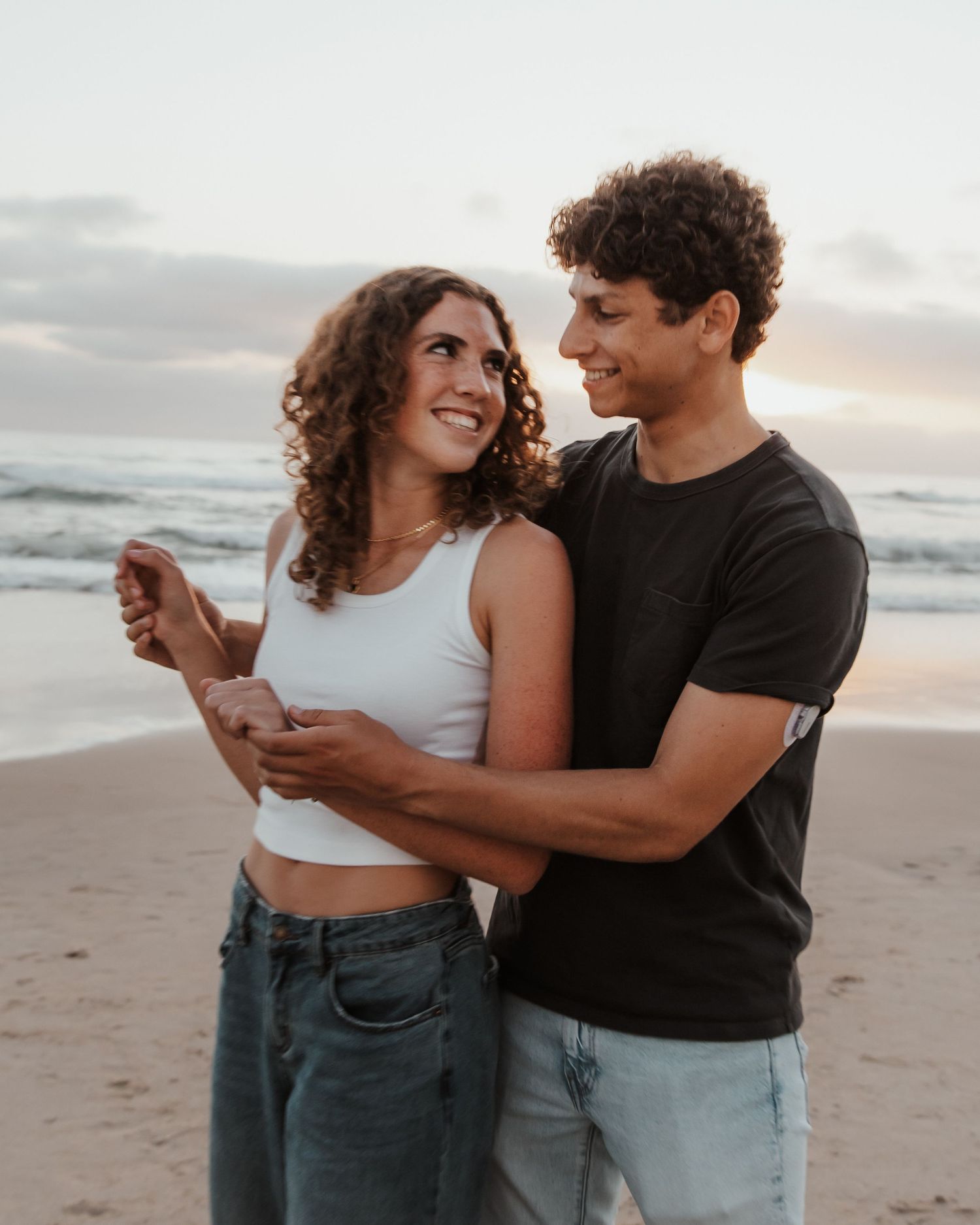 Couple in casual denim outfits dancing and embracing on a beach at sunset.