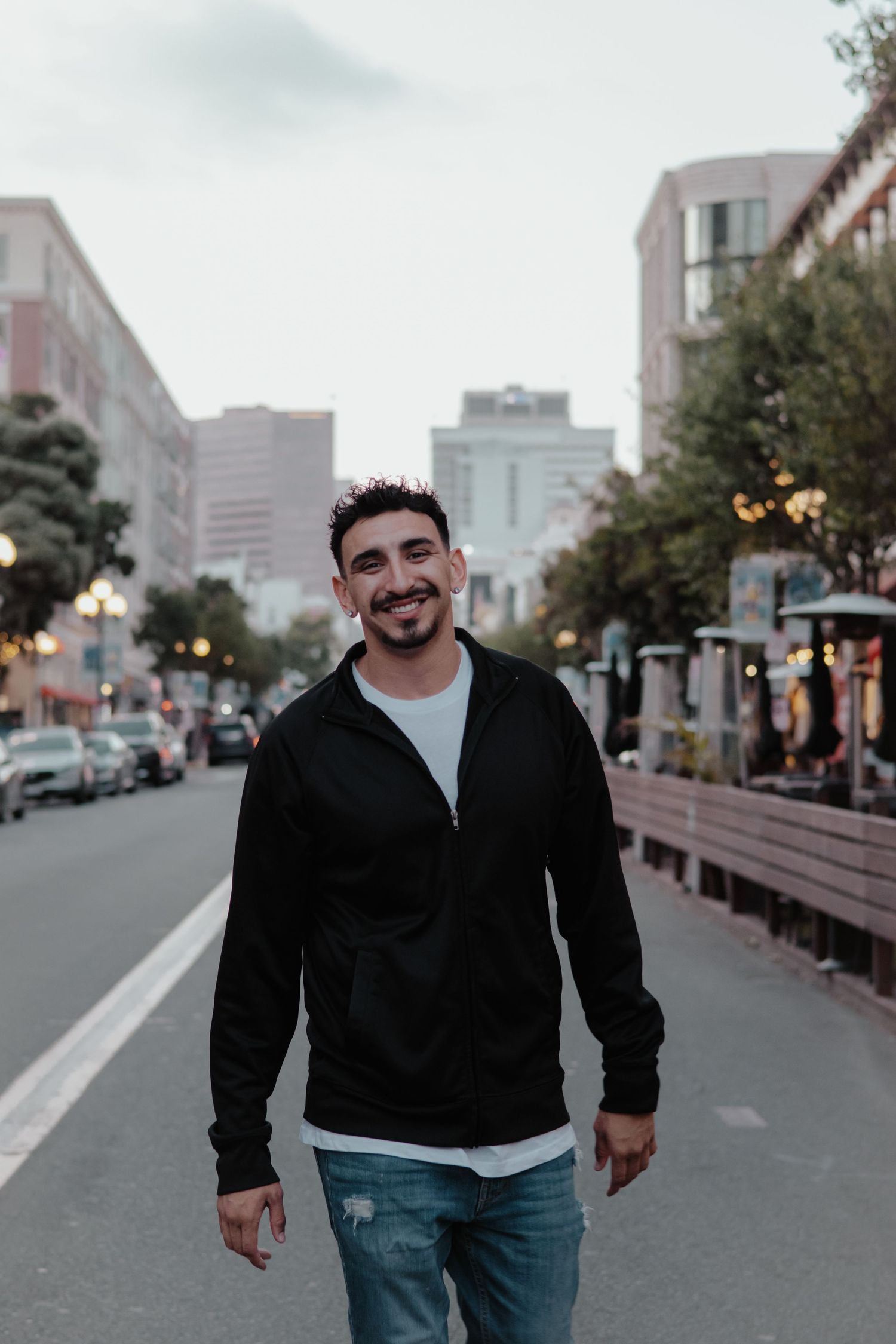 Person in black jacket and jeans walking down urban street at dusk.