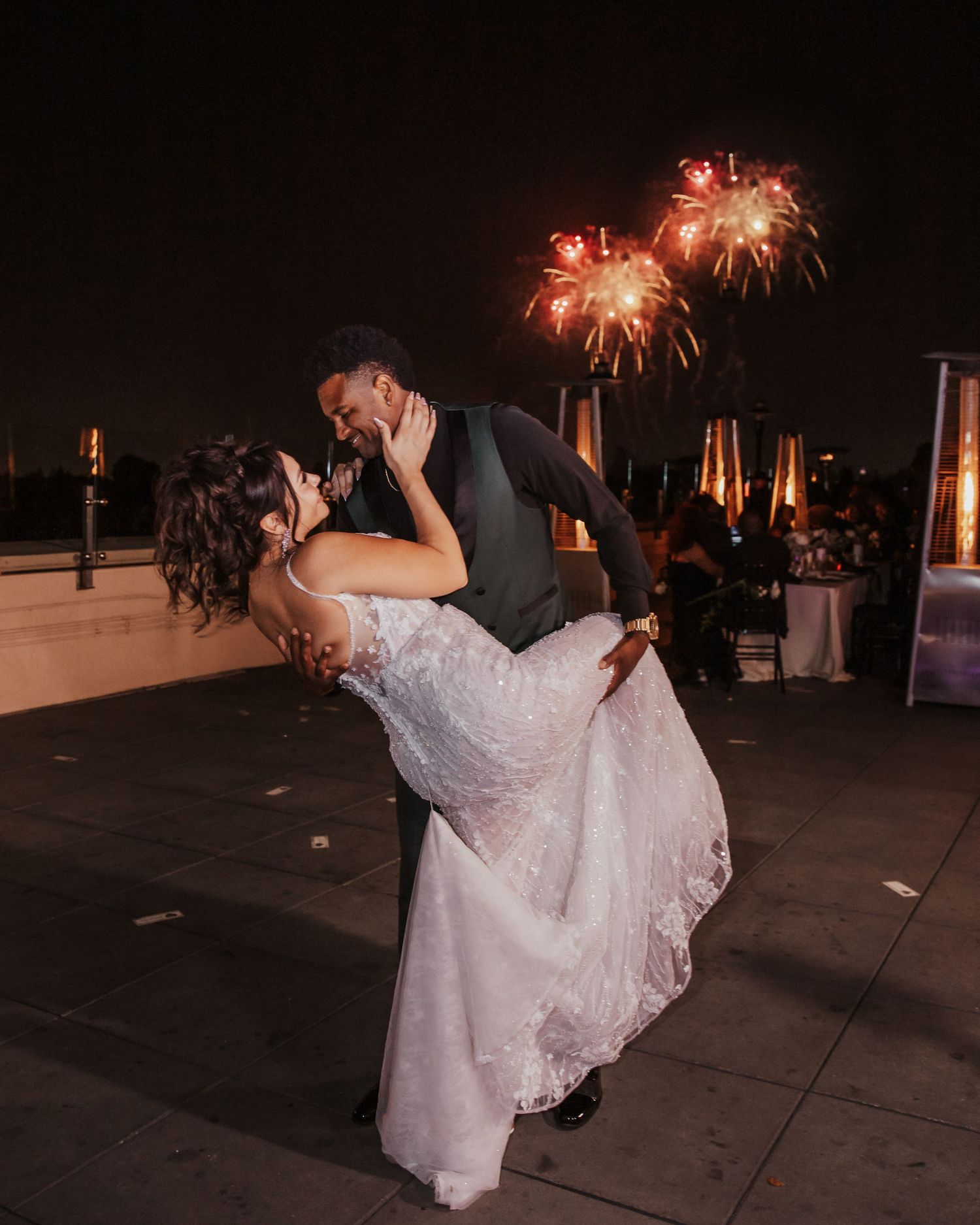 A romantic couple dancing and embracing under fireworks at an outdoor wedding celebration at night.