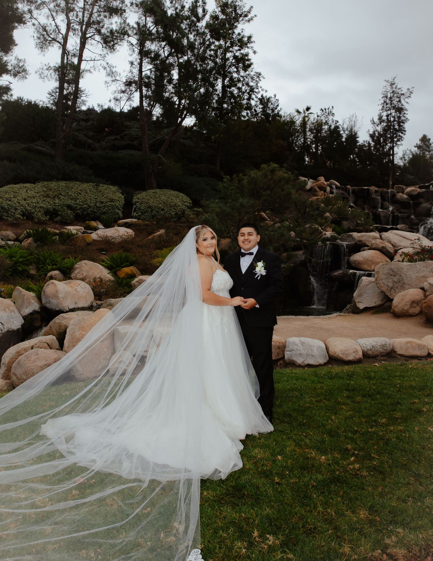 A wedding couple poses in front of a landscaped waterfall garden with rocks and trees on an overcast day.