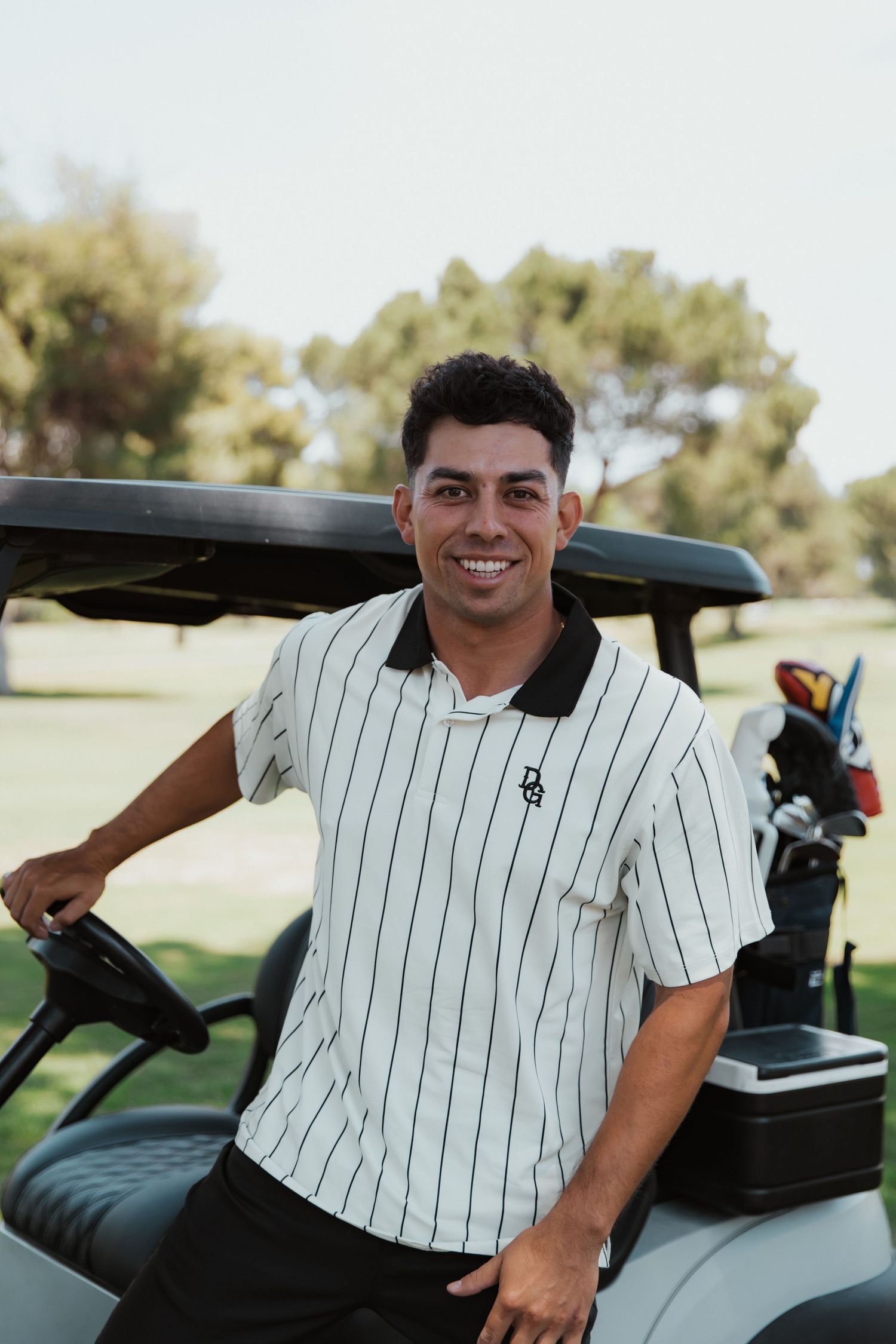 A golfer in a striped white polo shirt smiling next to a golf cart on a sunny course.