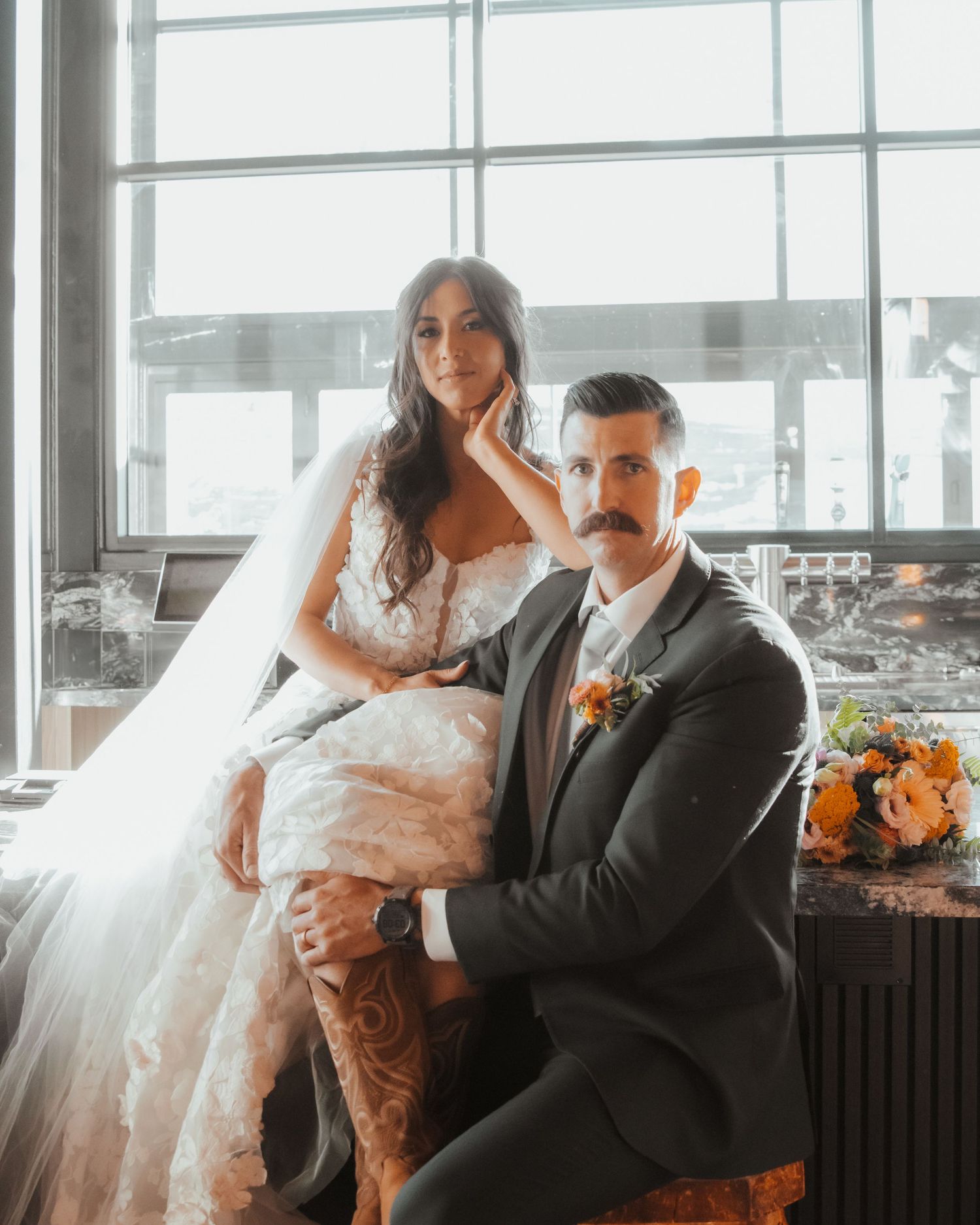 A couple poses with their dog in a modern venue with large windows and orange floral arrangements.