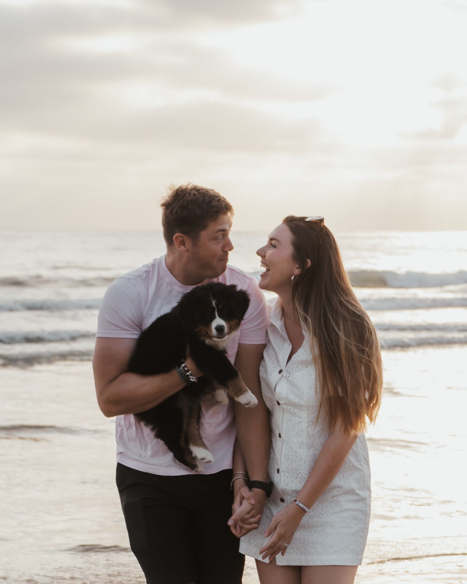 Couple at beach during sunset holding small black dog while sharing joyful moments.