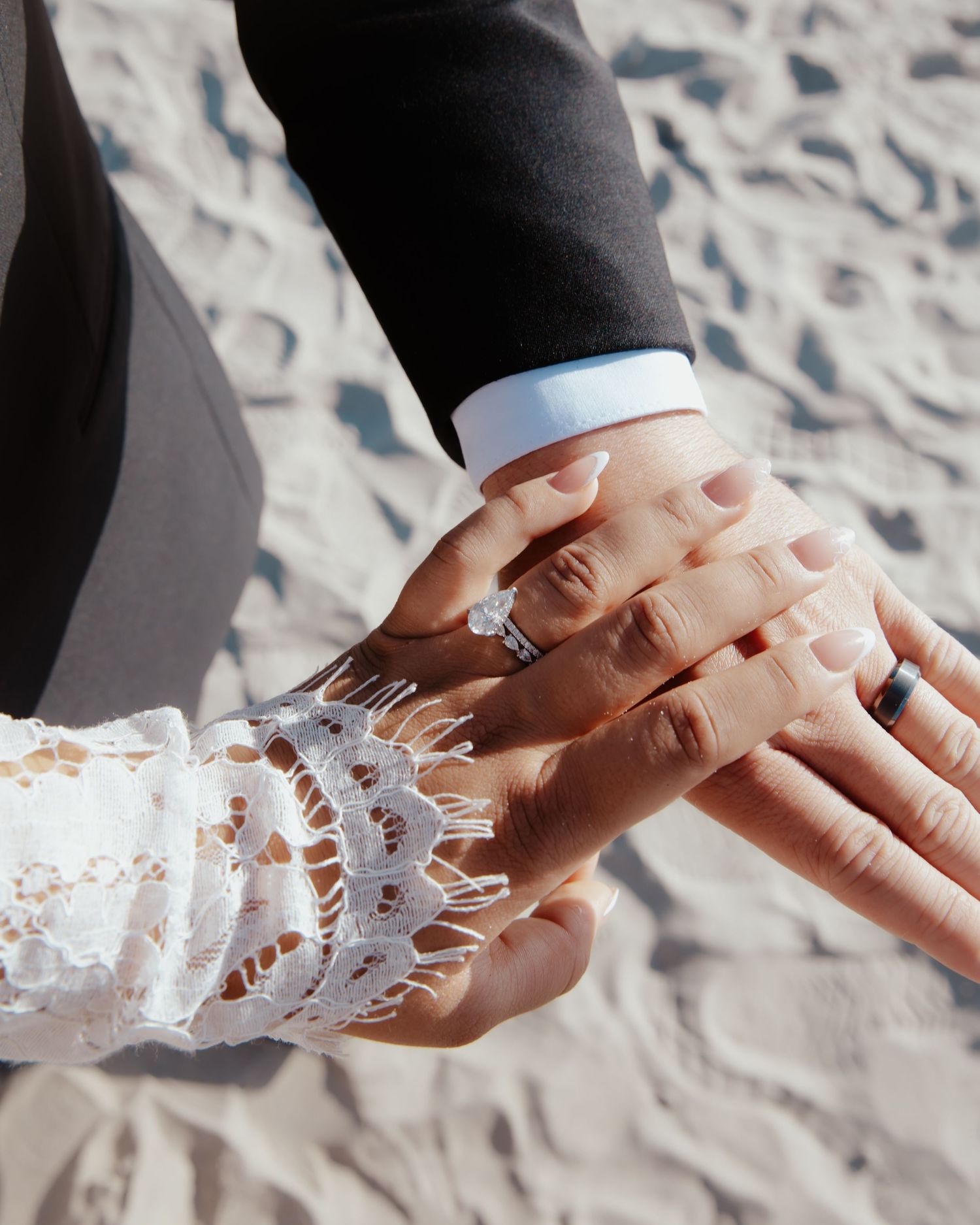 Close-up of hands with wedding rings touching on a sandy beach, showing lace sleeve detail.
