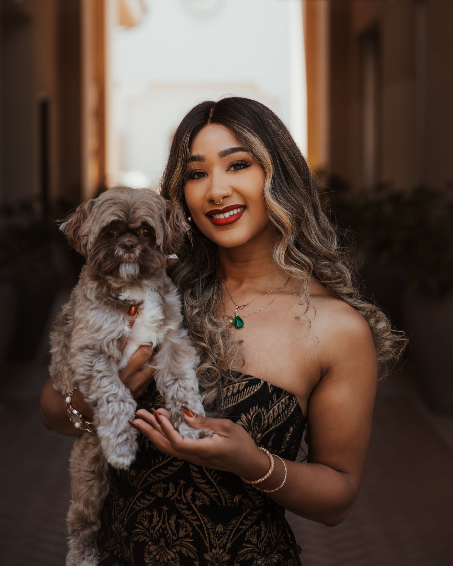 A person in an elegant black dress poses playfully with a fluffy gray cat in a warm-lit hallway.