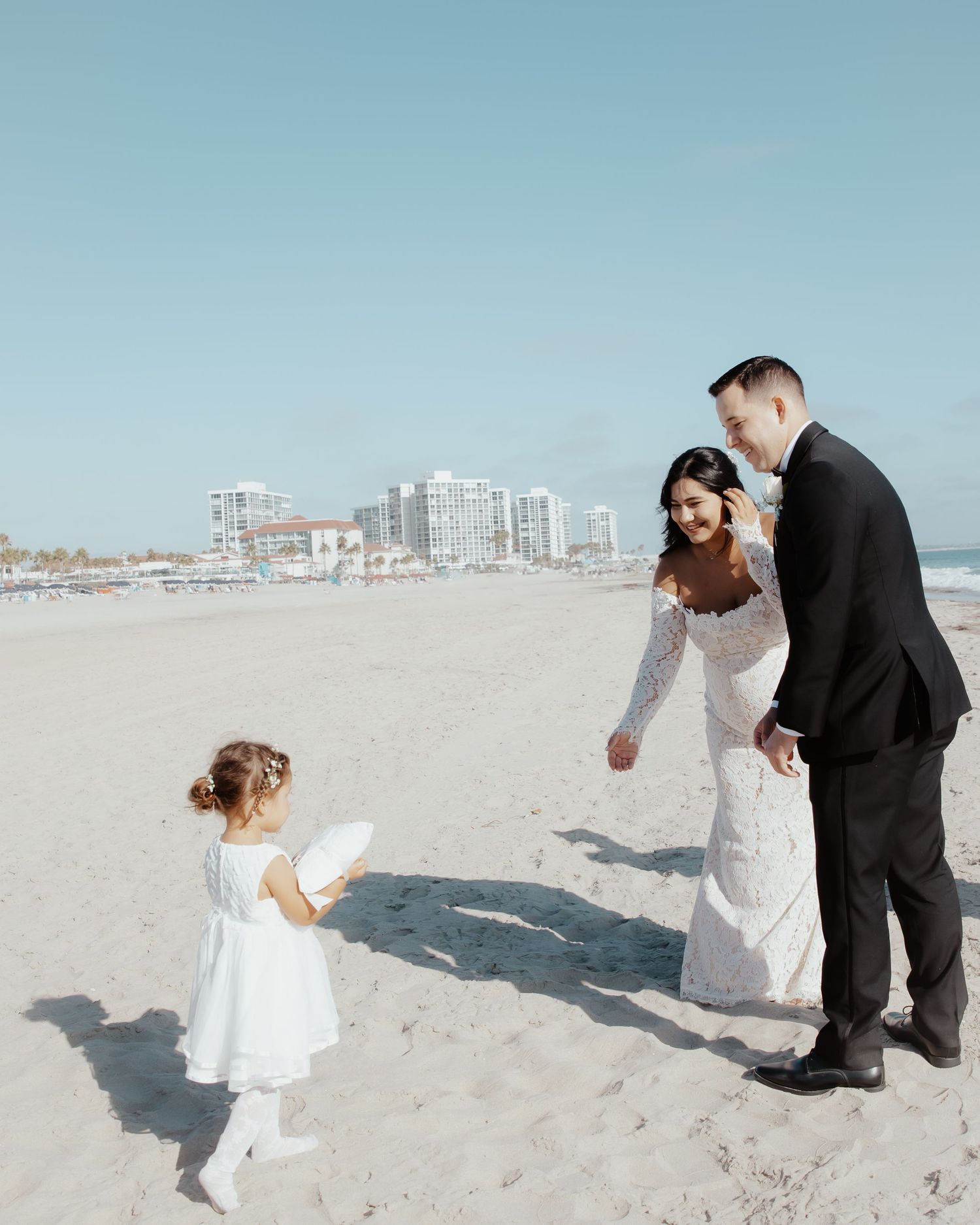 Wedding couple interacts with small child in white dress on sandy beach with buildings in background.
