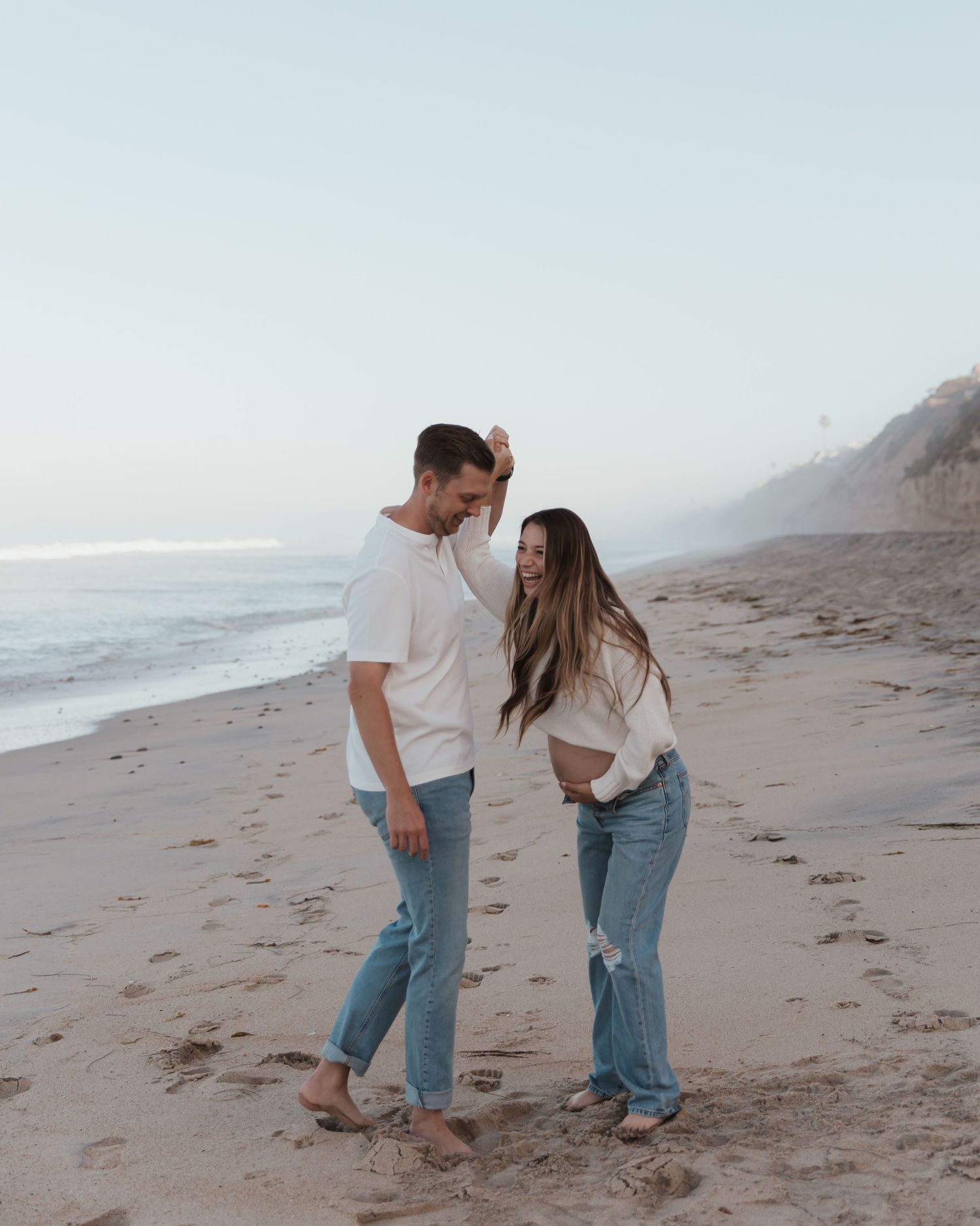 A couple shares a romantic moment while walking along a misty beach shoreline in casual denim outfits.