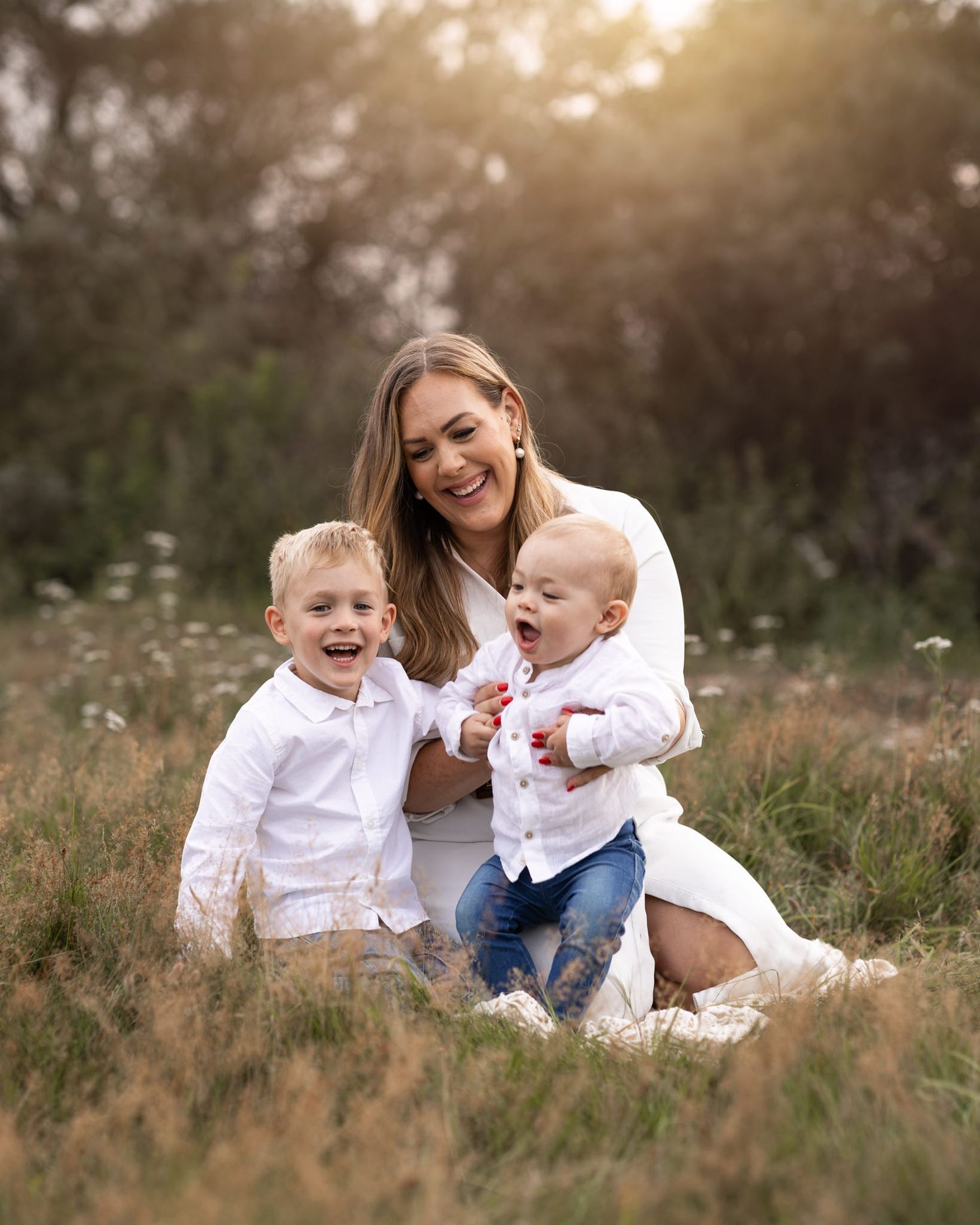 Moderskapsfotografering i Ängelholm. Mamma med två barn sitter höstgräs med solens gyllene strålar bakom dem. Alla är glada och ler