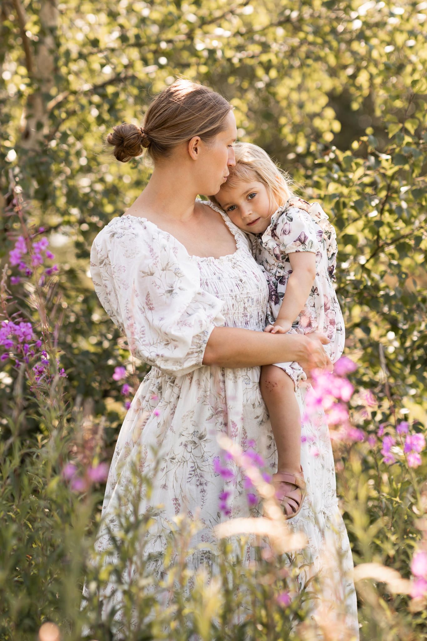 Moderskapsfotografering där en mamma håller sin dotter i famnen och pussar henne på pannan. En mysig stund utomhus en vacker sommardag.