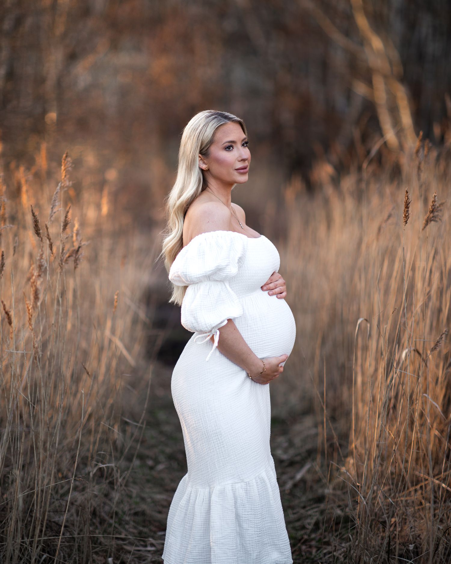 Gravidfotografering en höstdag bland vass. Vacker blond gravid kvinna omfamnar sin mage och tittar drömmande bort.