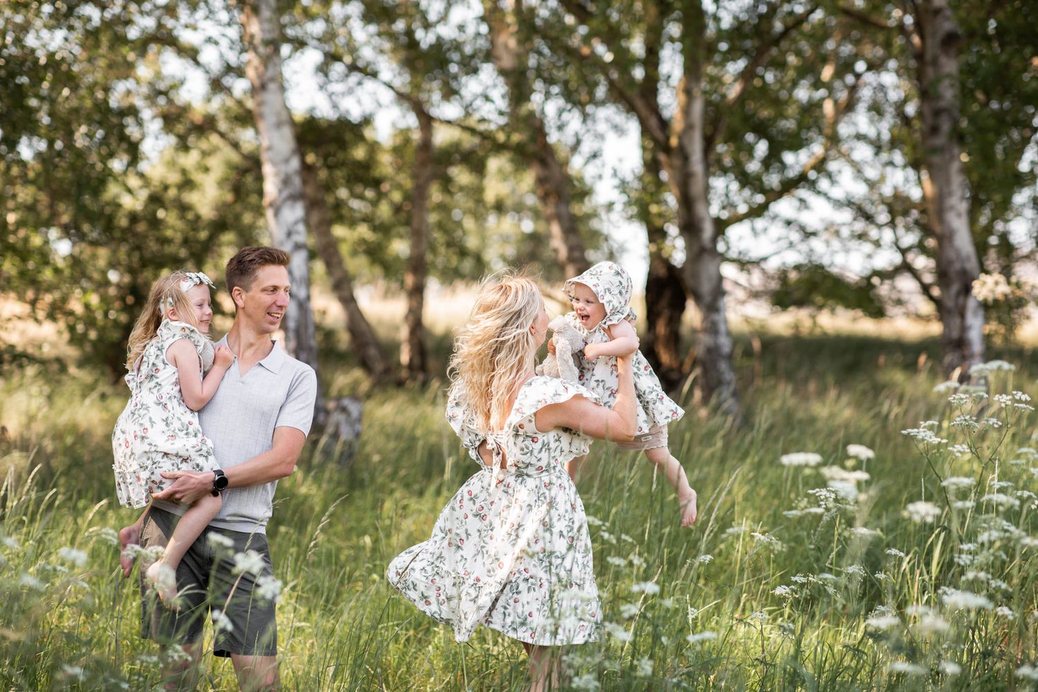 Lekfull familjefotografering där barnen är i föräldrarnas famn och busar på en vacker sommaräng med blommor och björkar i bakgrunden.
