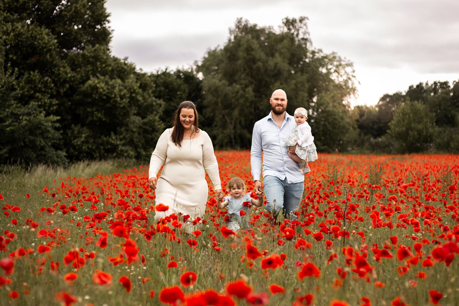 Familjefotografering i Skåne på ett vackert vallmofält. Familjen promenerar bland vallmon och håller varandra i händerna. Pappa bär på bebis