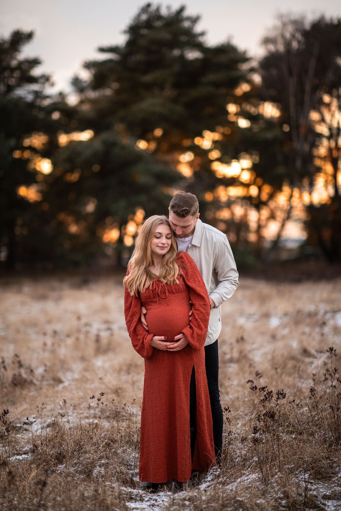 Gravidfotografering i vintergräs under solnedgång, hon i rostfärgad klänning och han bakom med armarna runt henne. Kärleksfull och naturlig.