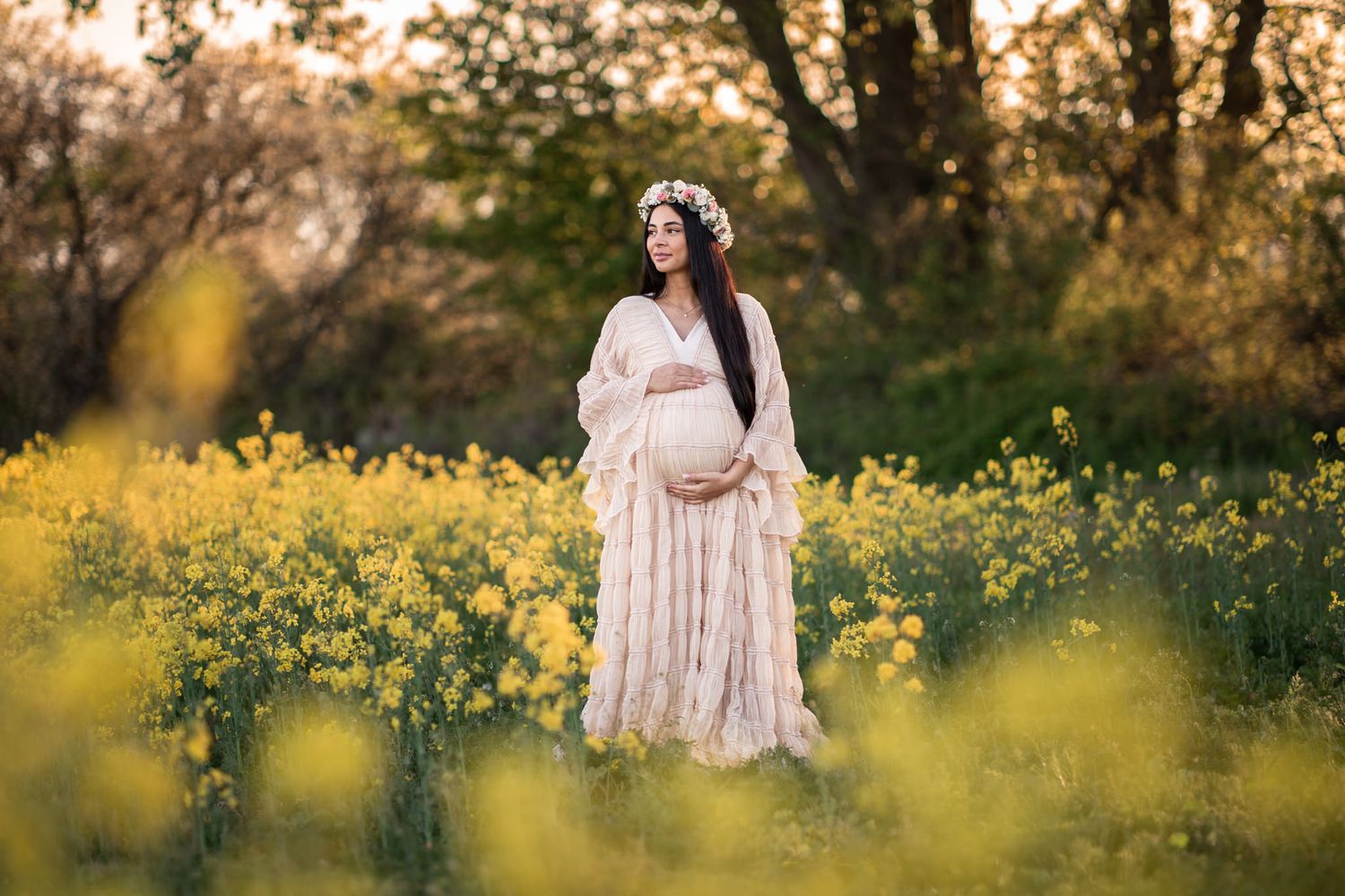 Gravidfotografering i Skåne, kvinna i ljus klänning och blomkrans står i ett gult rapsfält i solnedgången. Naturlig och varm känsla i bilden