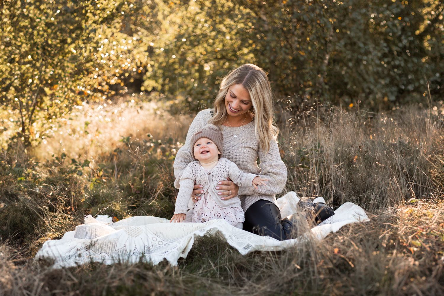 Moderskapsfotografering i Skåne med mamma och liten dotter. Sitter på filt i beigt höstgräs med solens gyllene strålar bakom dem. Båda ler