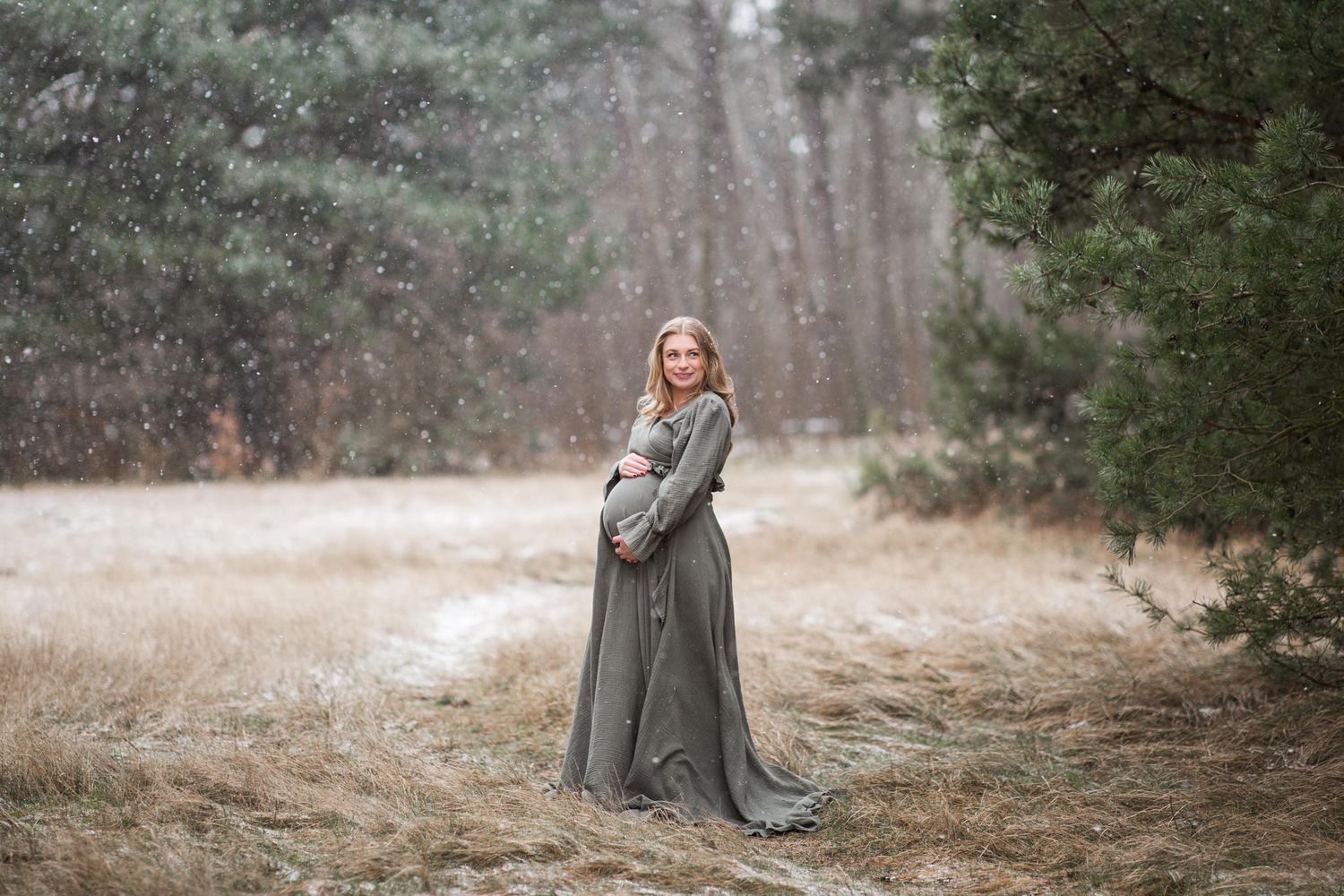 Gravidfotografering i Skånskt vinterlandskap. Beigt gräs och tallar i bakgrunden. Det faller snöflingor som börjat lägga sig på marken.