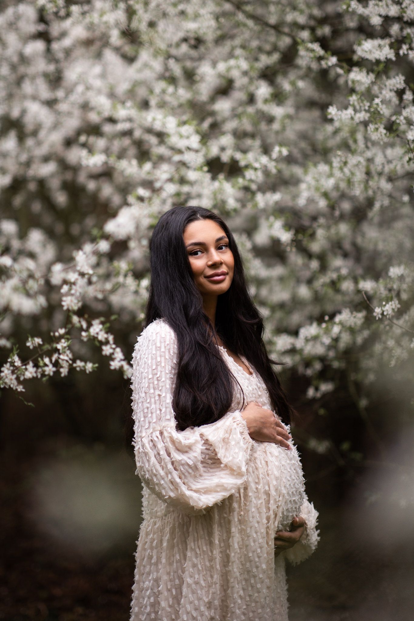 Vacker gravid kvinna som håller om sin mage och står utomhus bland mängder av blomstrande plommonträd. Hennes vita klänning matchar blommor.