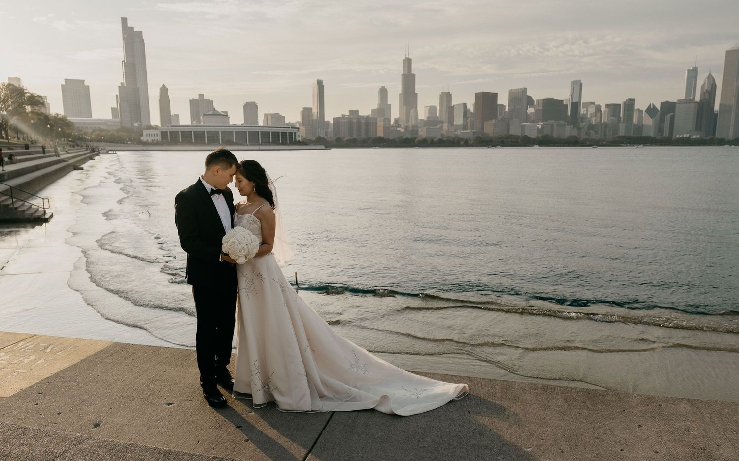 A bride and groom stand together by the lake Michigan with the Chicago skyline in the background, both wearing formal wedding attire.