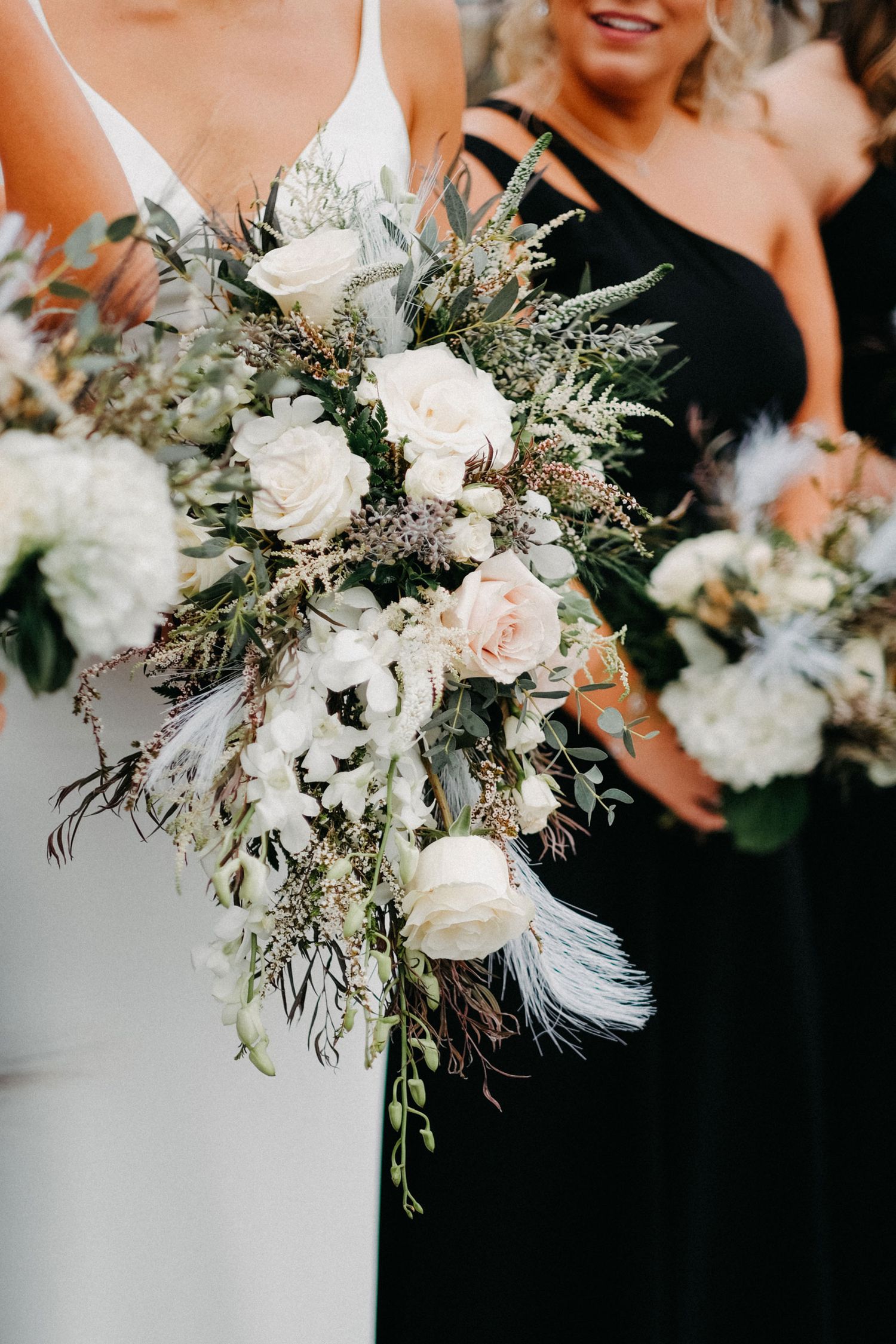 Bride holding a cascading bouquet of white and blush flowers with greenery and soft feathers alongside bridesmaids in black dresses; elegant