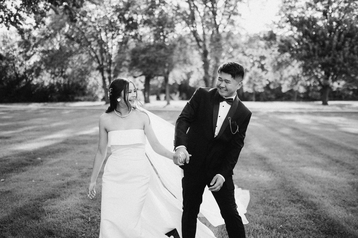 Bride and groom laughing while walking hand-in-hand across a sunlit lawn; candid black-and-white Chicago wedding photography.