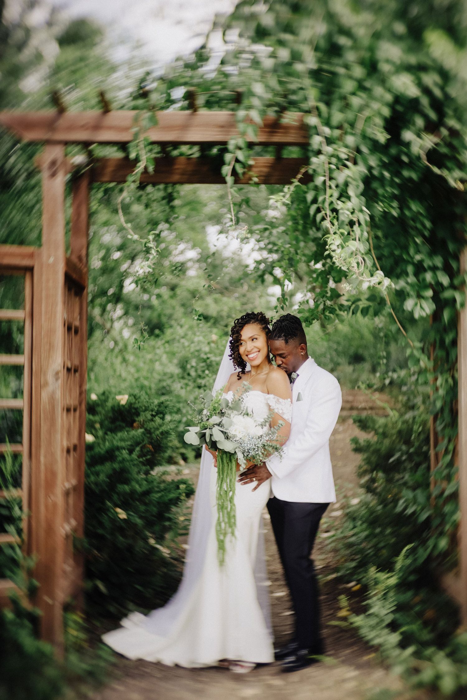 Bride and groom embracing under a wooden garden arbor draped in lush greenery, holding a cascading bouquet;romantic outdoor wedding portrait
