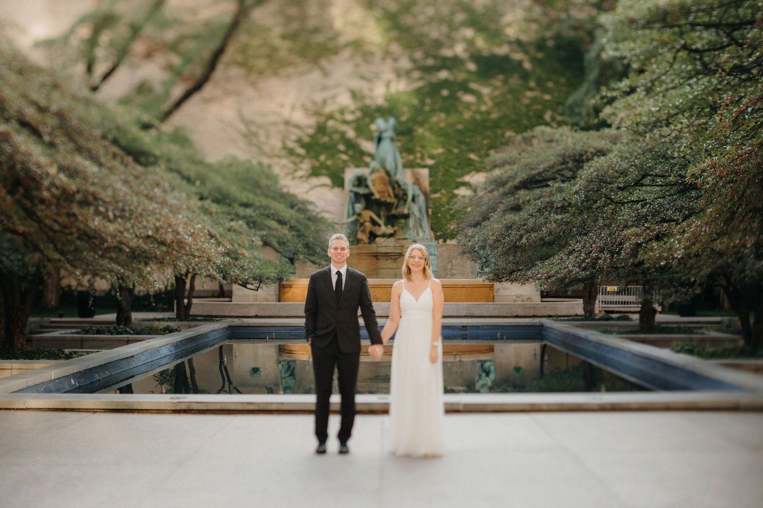 Bride and groom holding hands in front of a reflecting pool and statue near The Art Institute of Chicago; elegant garden wedding portrait.