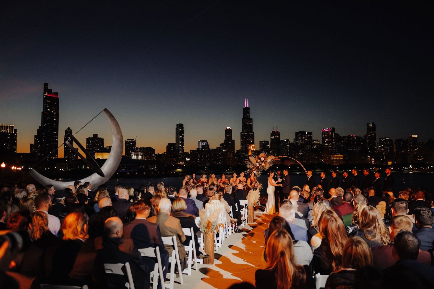 Outdoor wedding ceremony near Adler Planetarium at dusk, with Chicago skyline behind guests as couple stands under modern arch; cityscape we