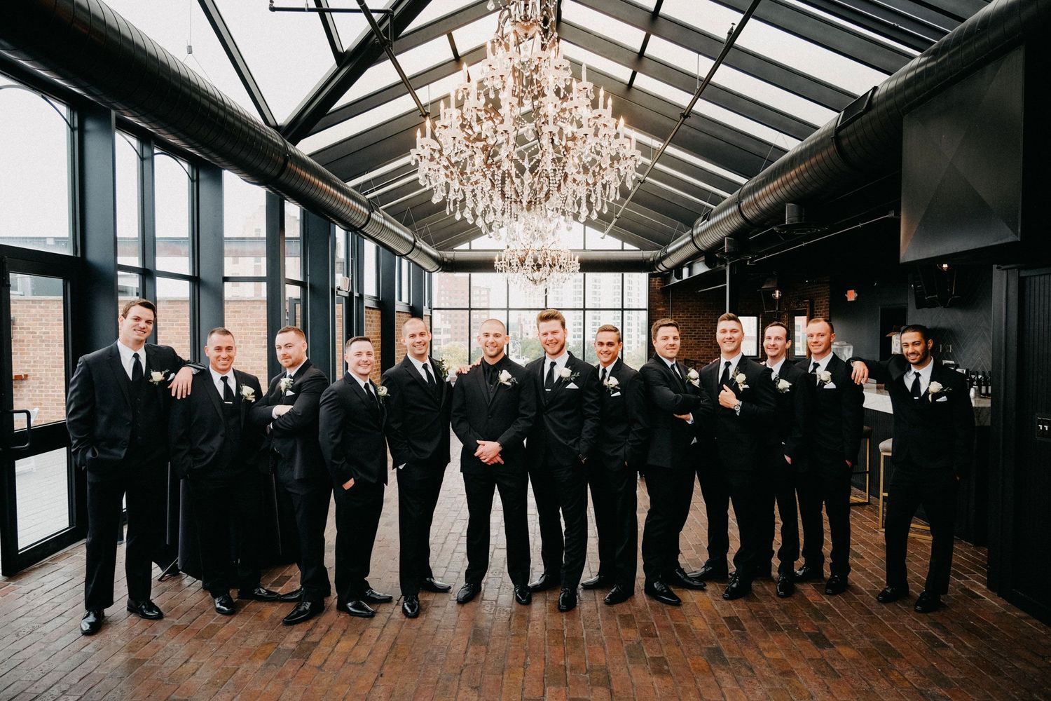 Group of men in black tuxedos posing in a modern glass-ceiling venue with chandeliers; luxury Chicago wedding party photo.