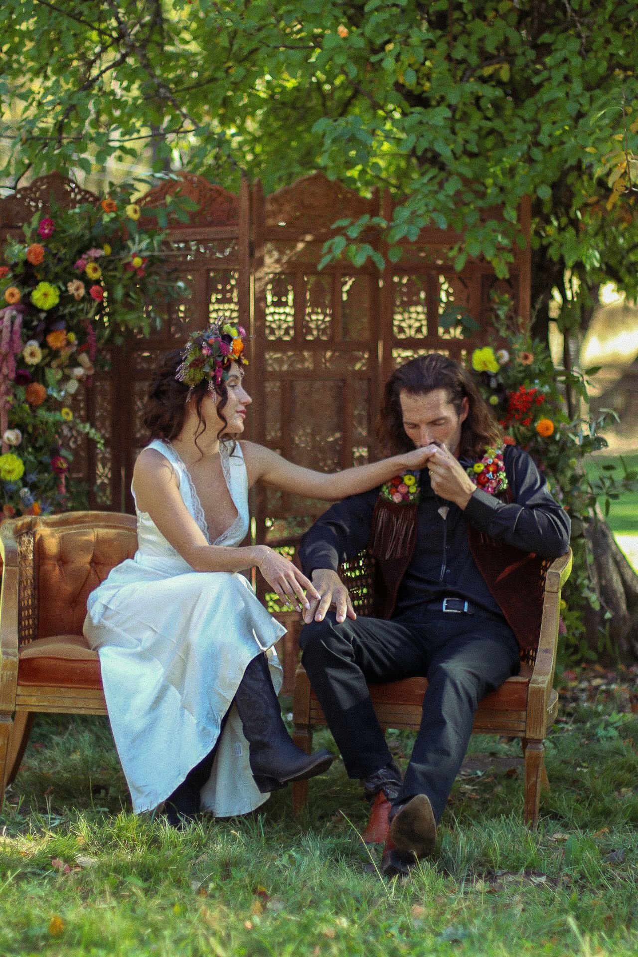 Bride in white dress and floral crown kisses her groom's hand while seated on vintage leather chairs at a boho outdoor garden wedding.