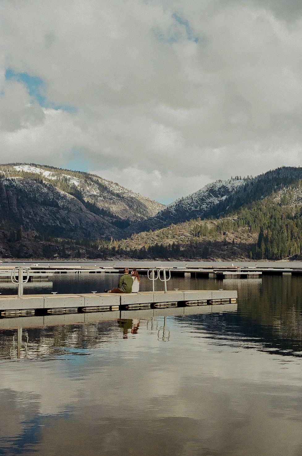 Two people sit together on a floating dock at a calm mountain lake, with snow-dusted hills and cloudy skies reflected in the water.