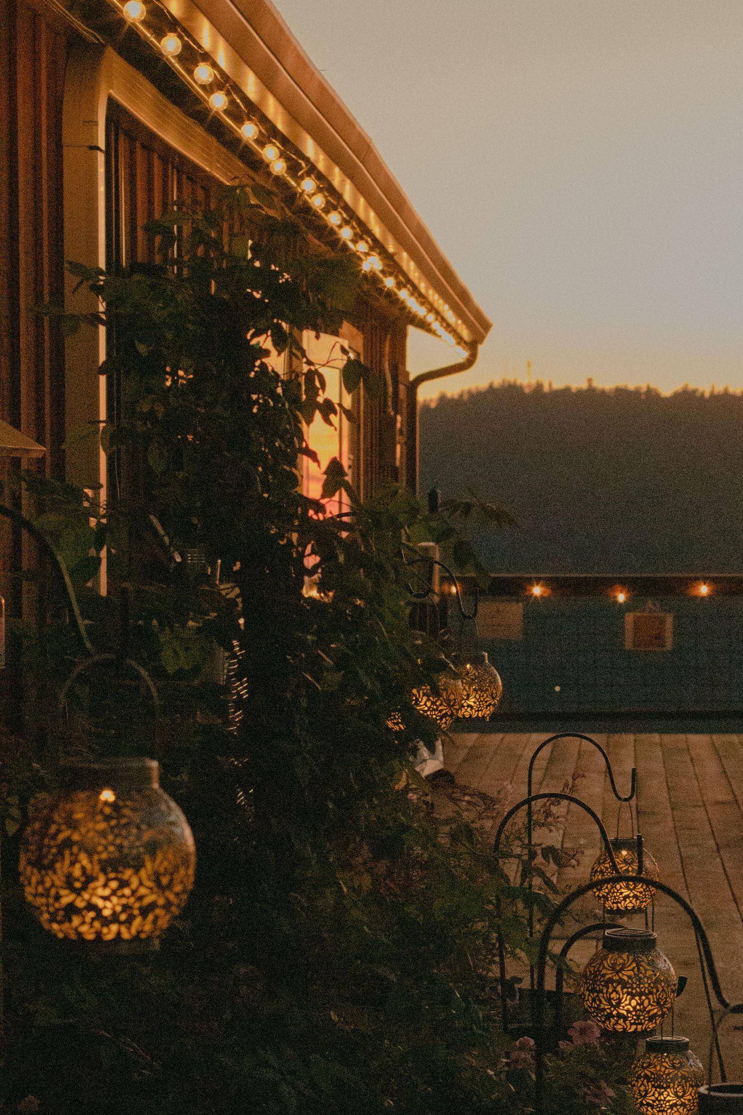 Glowing ornate lanterns hang among green vines beside a wooden building with string lights, overlooking a forested hillside at dusk.