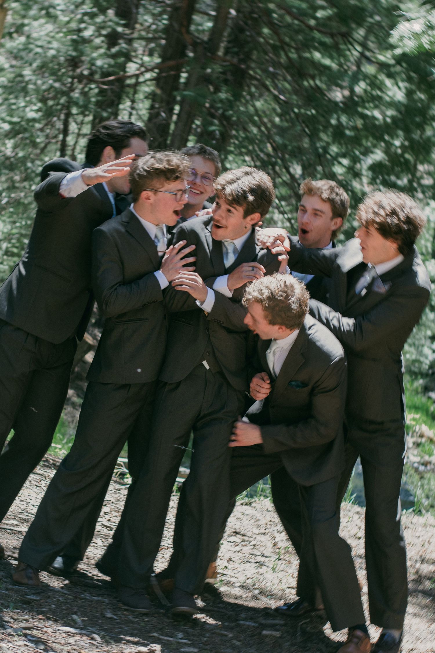 A group of groomsmen in dark suits laugh and playfully crowd together outdoors in a forest setting.
