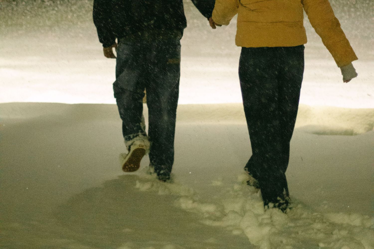 Two people in jeans and winter jackets walk hand in hand through deep snow at night as snowflakes fall around them.