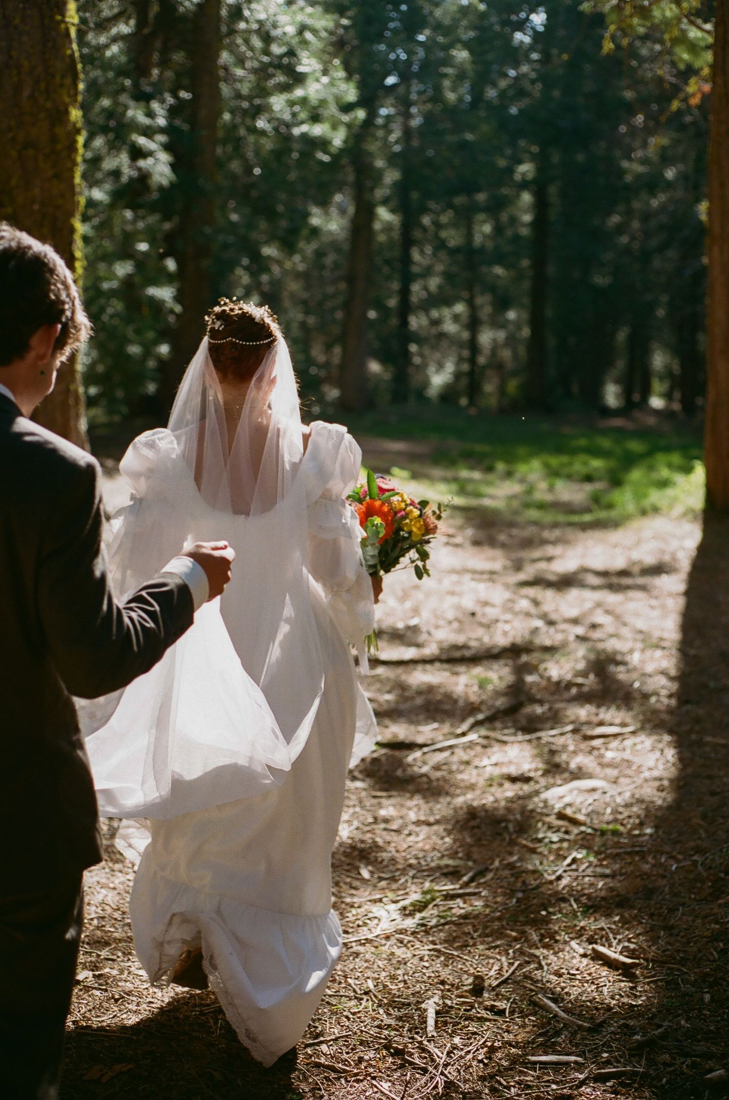 Bride in a white gown and veil carrying an orange bouquet walks down a sunlit forest path as a groom in a dark suit follows behind.