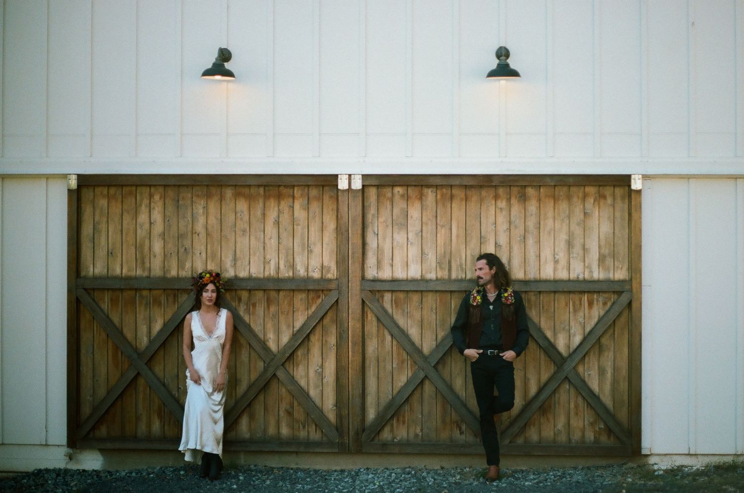 Two people stand on opposite sides of large rustic wooden barn doors with X-shaped trim under black gooseneck lights on a white wall.