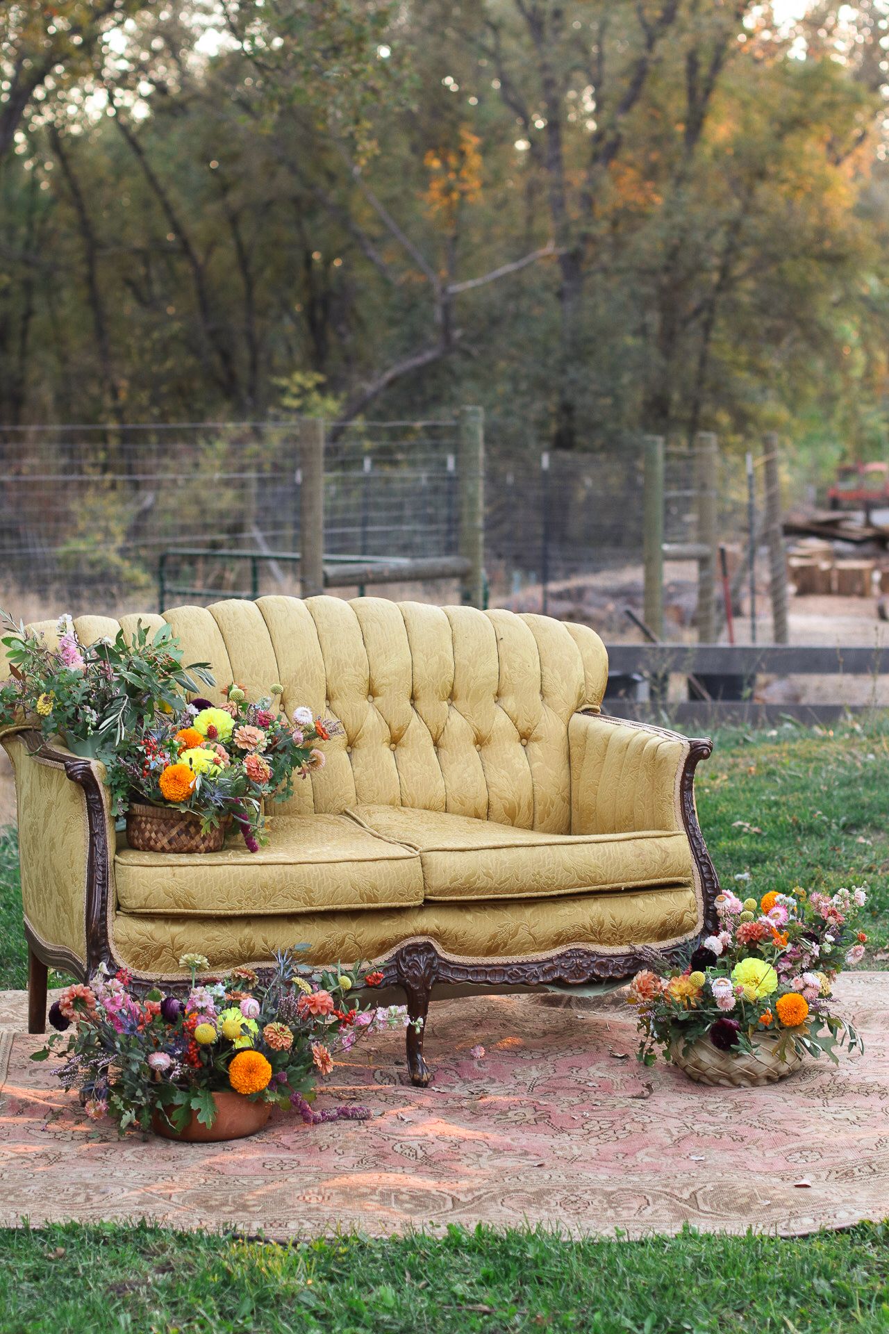 Vintage yellow tufted settee with ornate wood trim, surrounded by colorful floral arrangements on a rug outdoors near a rustic wire fence.
