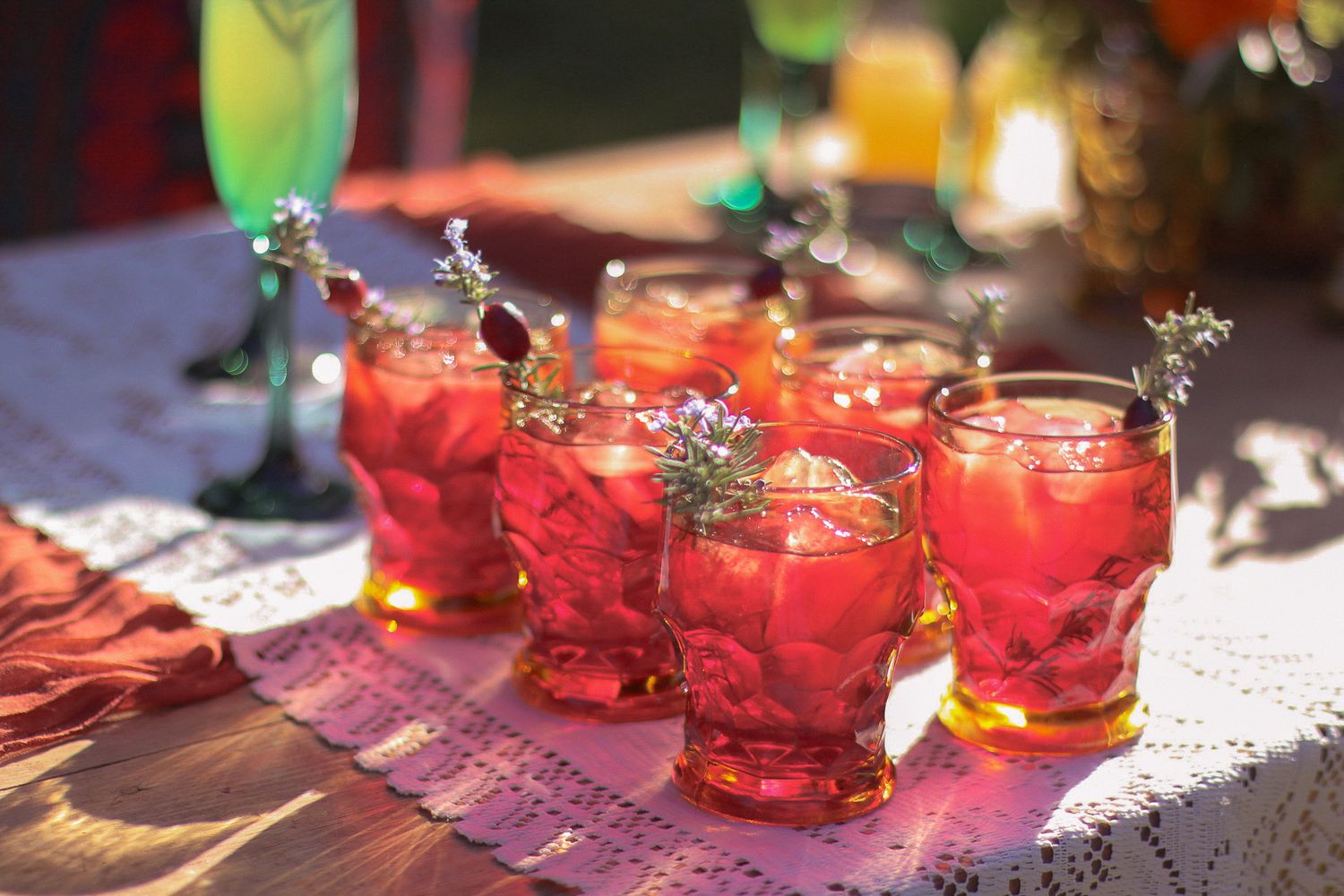 Red cocktails in glass tumblers garnished with herb sprigs sit on a lace-covered table in warm sunlight at an outdoor party.