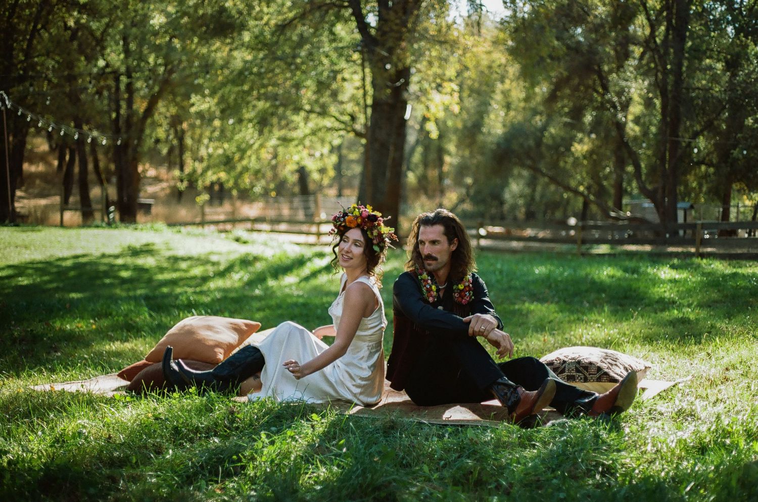 Couple sitting back to back on a picnic blanket in a sunny, tree-lined park, the woman in a white dress and flower crown.
