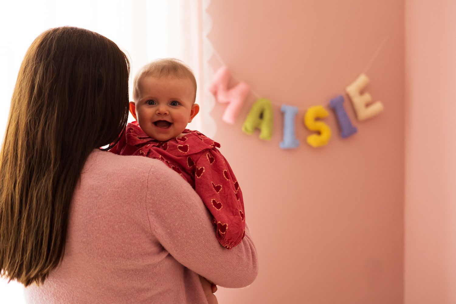 Mother holding baby girl beside a pink wall with name balloon letters, maisie name