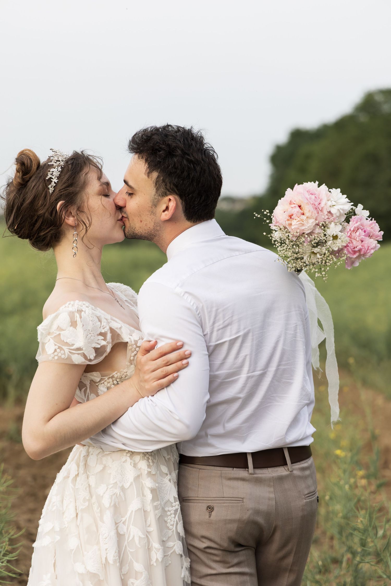 Groom kissing his partner's forehead while holding pink flowers in a field